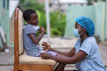 Cheerful African nurse in protective face mask seeing a child outdoor-concept on child health care in covid-19 pandemic Cheerful African nurse in protective face mask seeing a child outdoor-concept on child health care in covid-19 pandemic