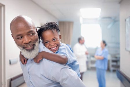 Bearded father giving piggyback to son at children's hospital. Male nurse and ill boy are in corridor. They are enjoying at hospital.